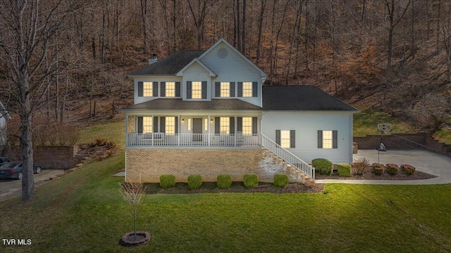 colonial-style house featuring a front yard, a forest view, covered porch, a chimney, and concrete driveway