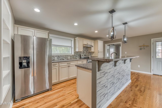 kitchen with light wood finished floors, backsplash, under cabinet range hood, stainless steel appliances, and a kitchen island with sink