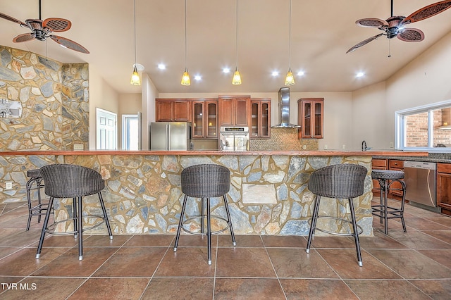 kitchen featuring wall chimney range hood, brown cabinetry, appliances with stainless steel finishes, and ceiling fan