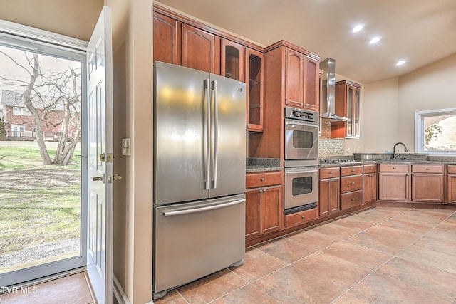 kitchen with glass insert cabinets, brown cabinets, stainless steel appliances, wall chimney exhaust hood, and a sink