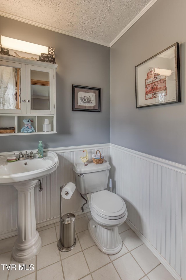 half bath featuring a wainscoted wall, toilet, ornamental molding, a textured ceiling, and tile patterned flooring