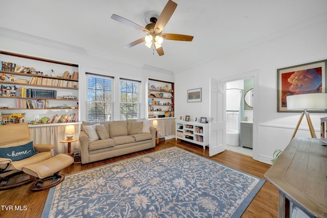 living room with built in features, wood finished floors, a wainscoted wall, ceiling fan, and crown molding