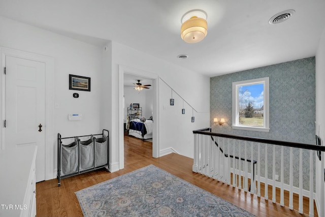 hall with wood finished floors, visible vents, baseboards, wallpapered walls, and an upstairs landing