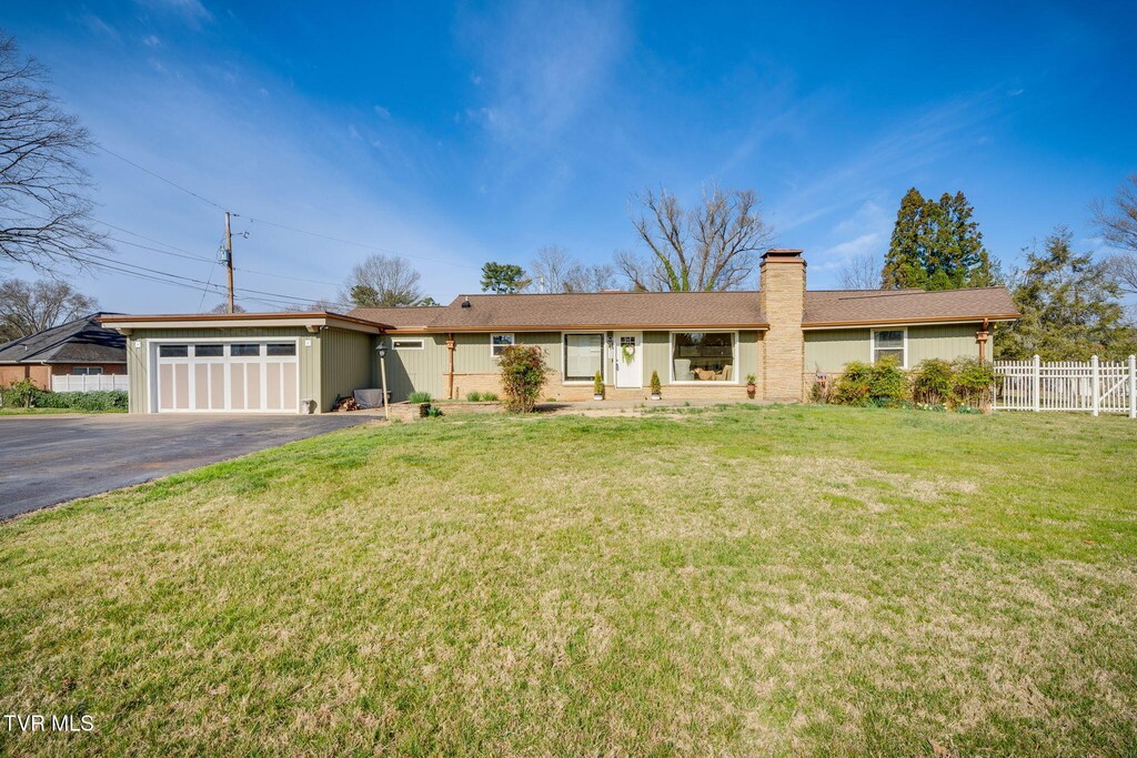 ranch-style home with driveway, a front yard, a chimney, an attached garage, and fence