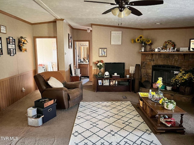 carpeted living room featuring crown molding, ceiling fan, a wainscoted wall, a stone fireplace, and a textured ceiling