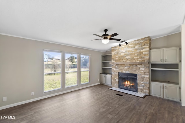 unfurnished living room featuring dark wood-type flooring, baseboards, crown molding, a stone fireplace, and a ceiling fan