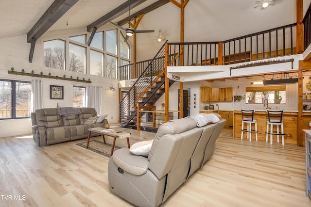 living room featuring stairs, light wood-style flooring, a ceiling fan, and beamed ceiling