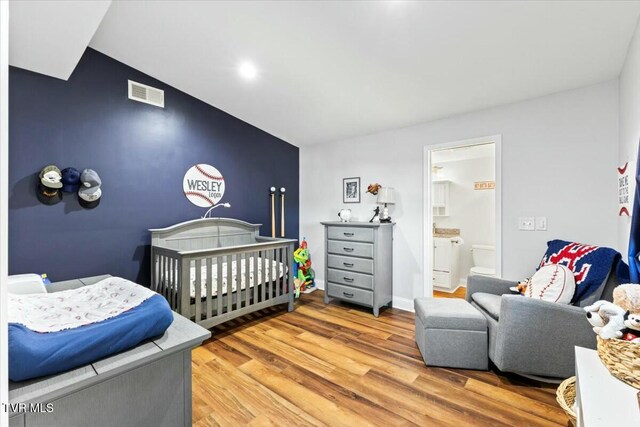 bedroom with visible vents, wood finished floors, a nursery area, baseboards, and an accent wall