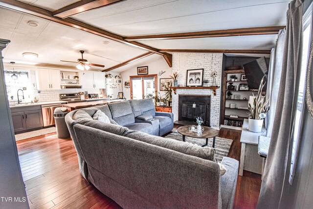 living area with a brick fireplace, lofted ceiling with beams, a ceiling fan, and wood finished floors