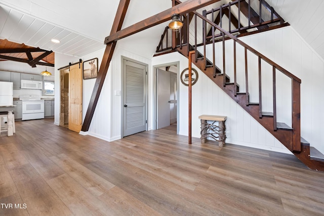 unfurnished living room featuring baseboards, stairway, a barn door, vaulted ceiling with beams, and wood finished floors