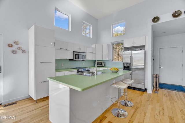 kitchen with a sink, a towering ceiling, white cabinets, light wood-style flooring, and appliances with stainless steel finishes