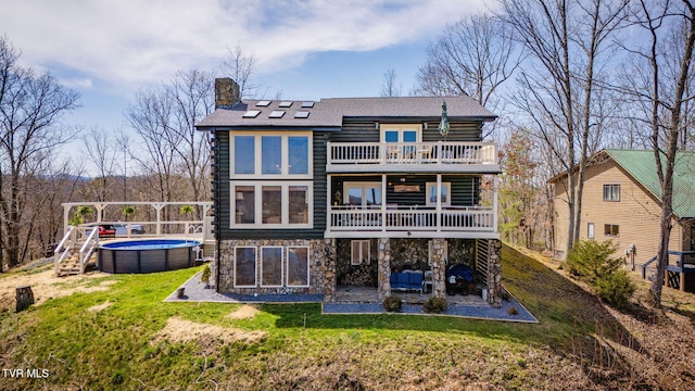 back of house featuring an outdoor pool, a lawn, stone siding, a balcony, and a chimney