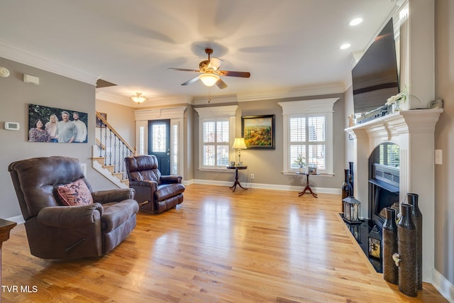 living area featuring ceiling fan, light wood-type flooring, a fireplace, crown molding, and stairs