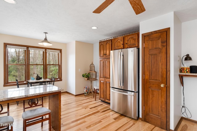 kitchen with light wood-type flooring, freestanding refrigerator, light stone countertops, brown cabinets, and ceiling fan