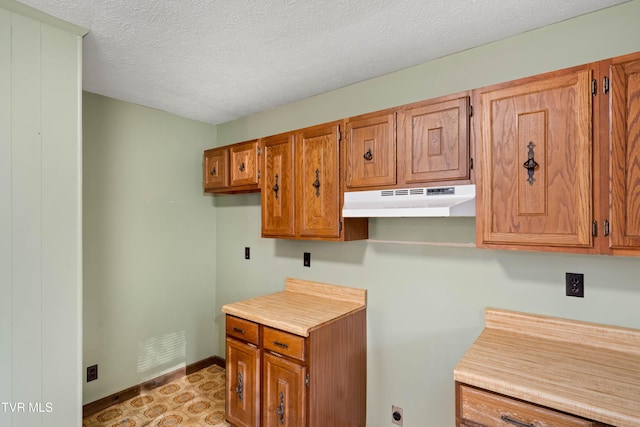 kitchen with baseboards, light countertops, a textured ceiling, under cabinet range hood, and brown cabinets