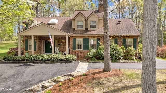 cape cod home featuring brick siding, a shingled roof, a chimney, and covered porch