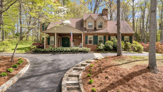 cape cod home featuring brick siding, driveway, a chimney, and roof with shingles