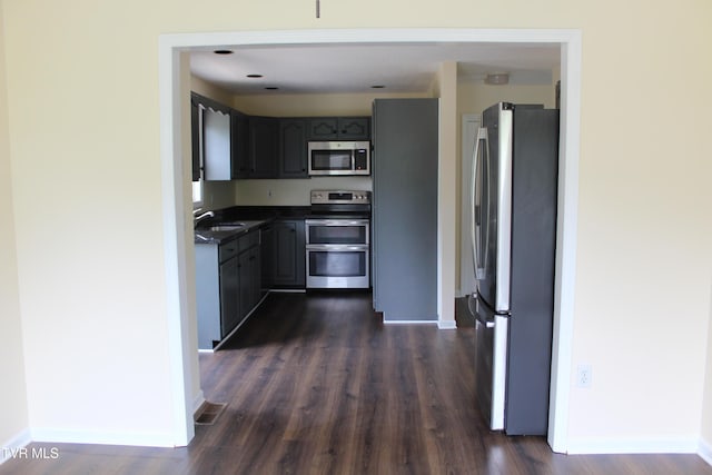 kitchen with stainless steel appliances, a sink, dark countertops, baseboards, and dark wood-type flooring
