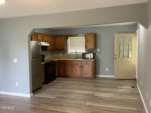 kitchen with light wood finished floors, a sink, brown cabinets, under cabinet range hood, and black appliances