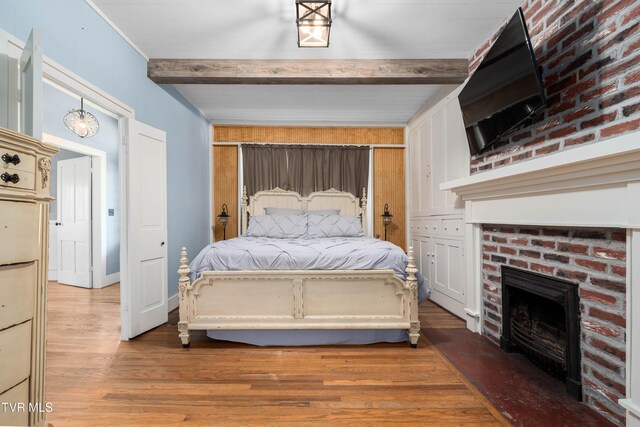 bedroom with beam ceiling, light wood-type flooring, and a brick fireplace