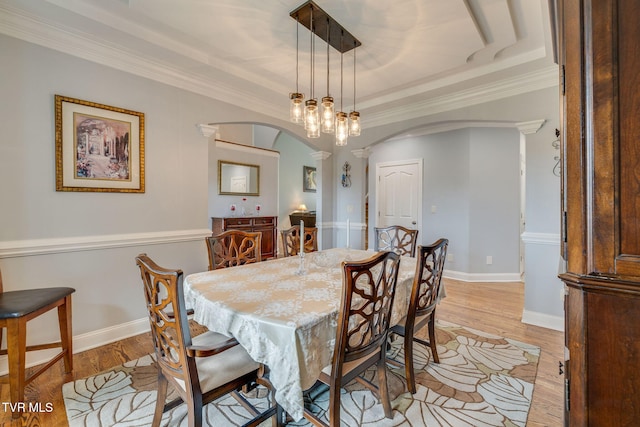 dining space with arched walkways, ornate columns, ornamental molding, light wood-type flooring, and a raised ceiling