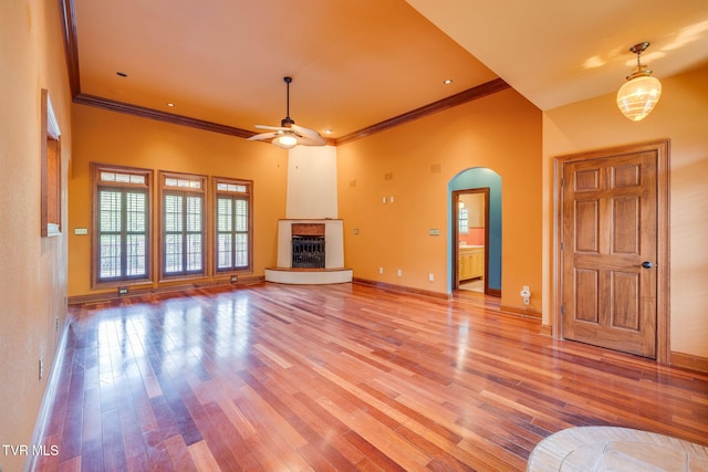 unfurnished living room featuring a fireplace with raised hearth, ceiling fan, crown molding, arched walkways, and wood finished floors