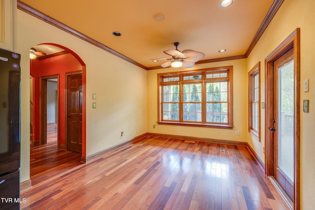 empty room with crown molding, light wood-style floors, baseboards, arched walkways, and a ceiling fan