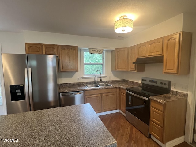 kitchen featuring stainless steel appliances, under cabinet range hood, dark countertops, dark wood-style flooring, and a sink