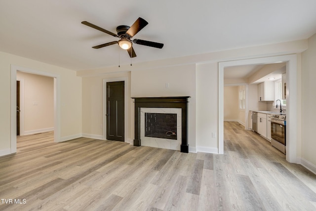 unfurnished living room with a sink, a tiled fireplace, ceiling fan, light wood-type flooring, and baseboards