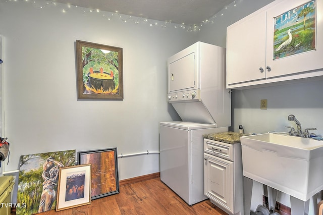 laundry room featuring cabinet space, stacked washer / drying machine, a sink, baseboards, and wood finished floors