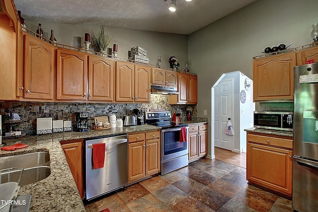 kitchen featuring appliances with stainless steel finishes, stone finish floor, stone counters, tasteful backsplash, and under cabinet range hood