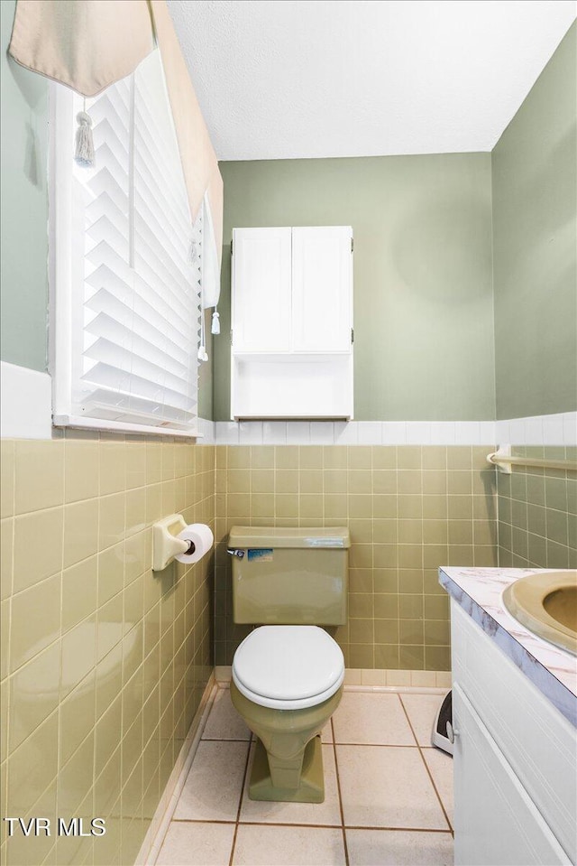 half bath with vanity, toilet, tile patterned floors, a wainscoted wall, and tile walls