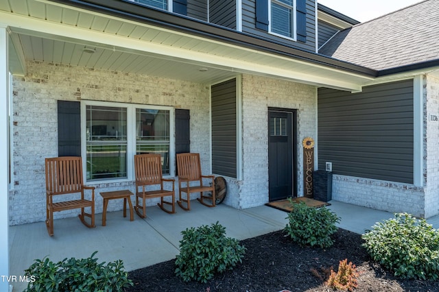 entrance to property featuring brick siding, a shingled roof, and covered porch