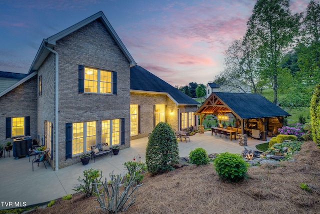 back of house at dusk featuring brick siding, a gazebo, cooling unit, an outdoor living space with a fire pit, and a patio