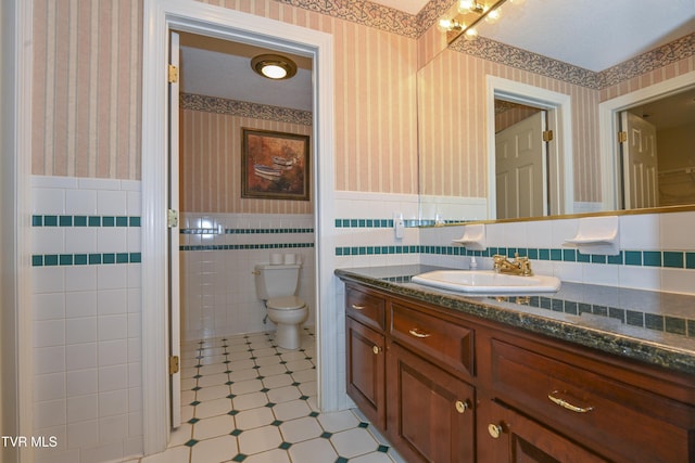 bathroom with tile walls, wainscoting, vanity, toilet, and wallpapered walls