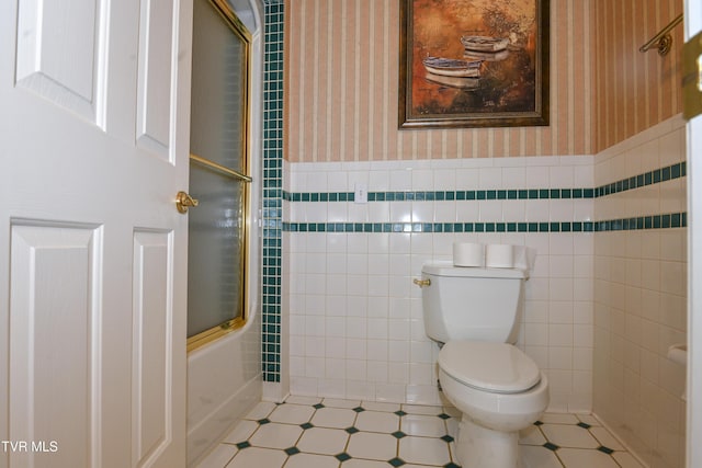 bathroom featuring wainscoting, toilet, tile walls, wallpapered walls, and bath / shower combo with glass door
