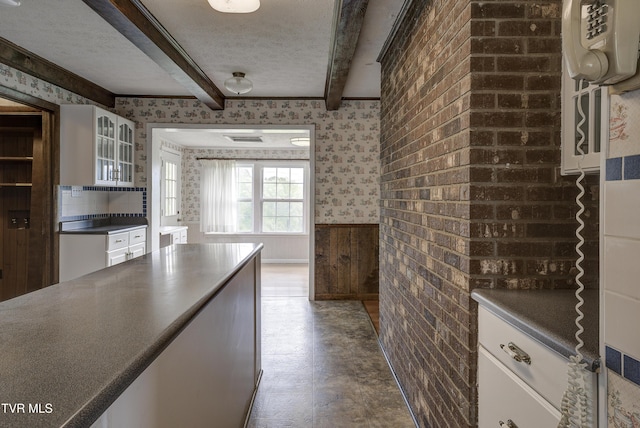 kitchen with wainscoting, wallpapered walls, white cabinetry, beam ceiling, and dark countertops