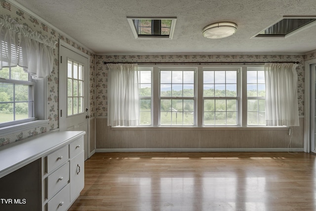 unfurnished dining area featuring wallpapered walls, light wood finished floors, wainscoting, a textured ceiling, and baseboards