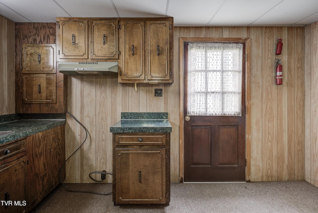 kitchen with under cabinet range hood, dark countertops, carpet floors, wood walls, and brown cabinetry