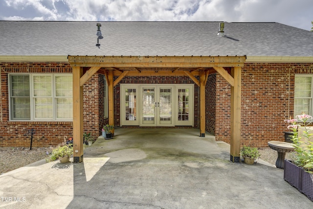 doorway to property featuring a shingled roof, french doors, brick siding, and a patio