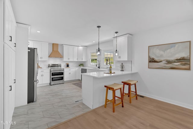 kitchen featuring appliances with stainless steel finishes, a peninsula, wall chimney exhaust hood, a kitchen bar, and white cabinetry