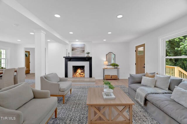 living area featuring recessed lighting, healthy amount of natural light, a warm lit fireplace, decorative columns, and light wood-style flooring