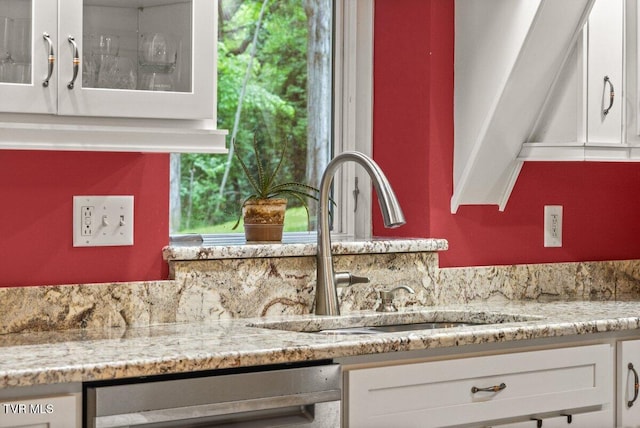 detailed view of a sink, dishwashing machine, white cabinetry, light stone countertops, and glass insert cabinets