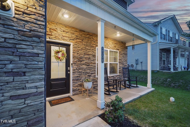 exterior entry at dusk featuring stone siding, a porch, and a lawn