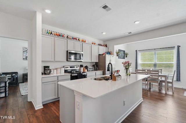 kitchen featuring appliances with stainless steel finishes, a sink, a kitchen island with sink, dark wood-style floors, and light countertops