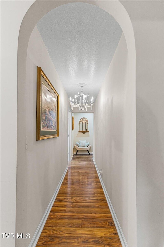hallway with wood finished floors, a textured ceiling, a chandelier, baseboards, and arched walkways
