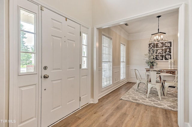 foyer featuring a chandelier, light wood-style flooring, a wainscoted wall, crown molding, and a decorative wall