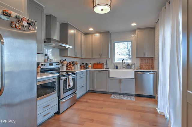 kitchen with stainless steel appliances, wall chimney range hood, gray cabinetry, a sink, and light wood-style flooring