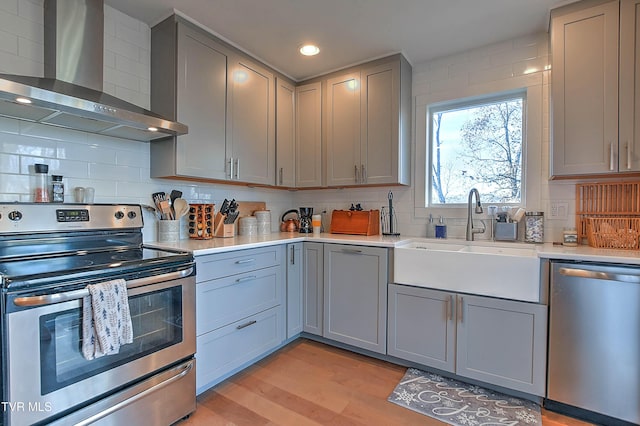 kitchen featuring stainless steel appliances, wall chimney exhaust hood, gray cabinets, a sink, and light countertops