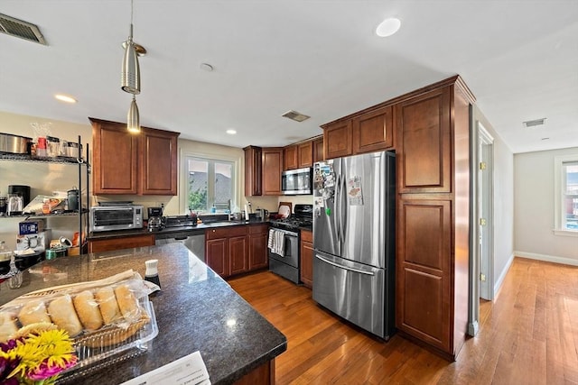 kitchen with appliances with stainless steel finishes, a sink, wood finished floors, hanging light fixtures, and dark stone counters
